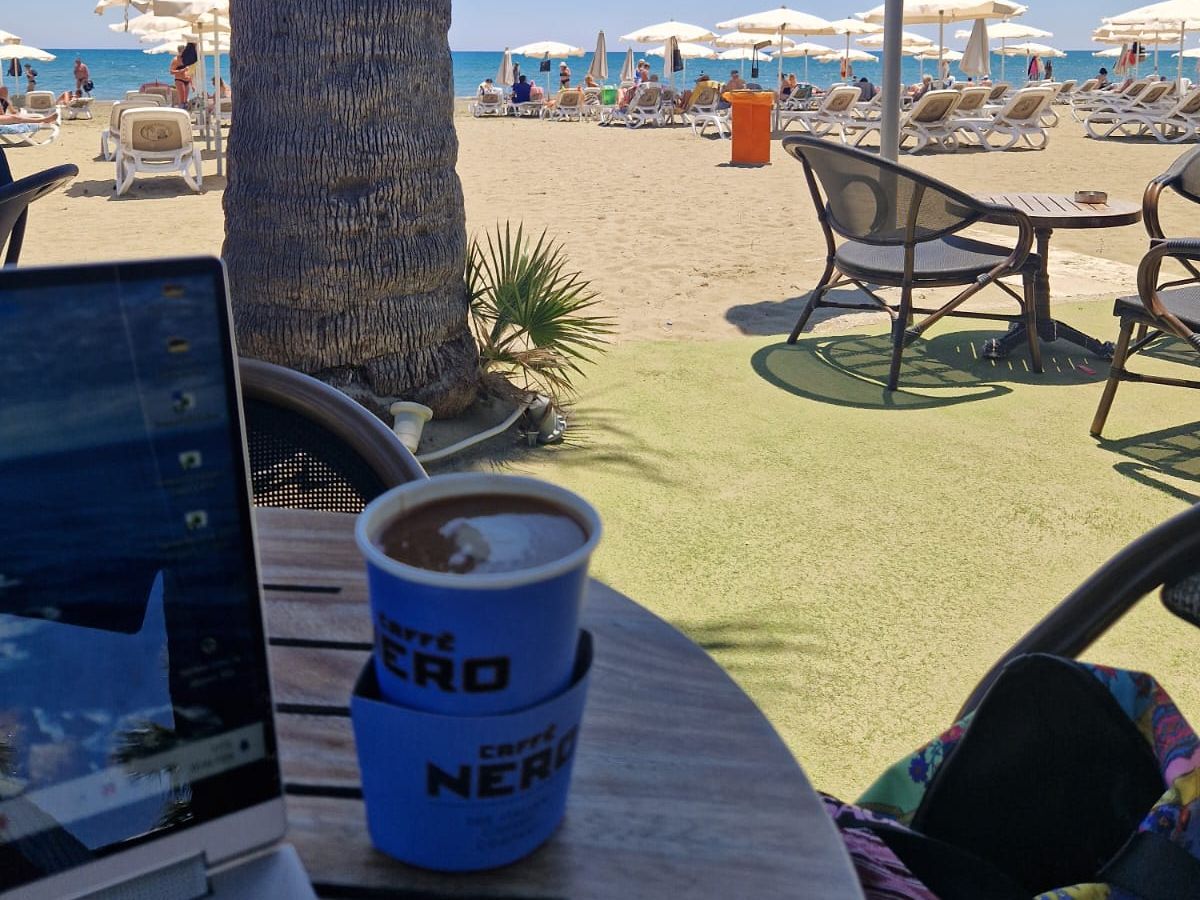 Laptop auf einem Tisch am Strand von Larnaka, dahinter Sonnenschirme und Meer